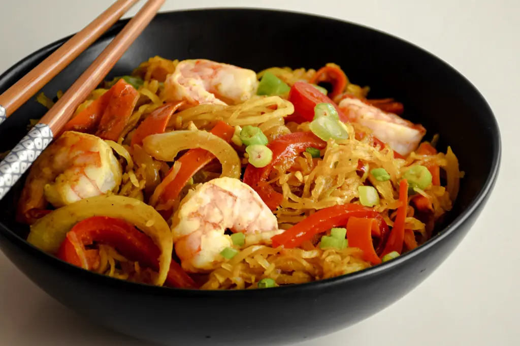 Three‑quarter angle close‑up shot of Singapore curry noodles served in a black bowl, featuring yellow noodles, prawns, sliced vegetables, and curry sauce, garnished with green onions.