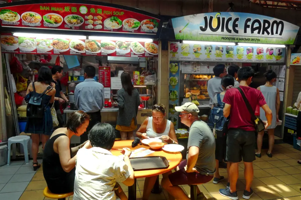 Wide-angle documentary-style shot of diners eating at a crowded hawker centre, with food stalls, menus, and customers lining up in the background.