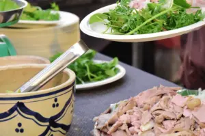 A food vendor preparing pho, holding a plate of fresh green herbs over a table filled with bowls and thinly sliced cooked meats.