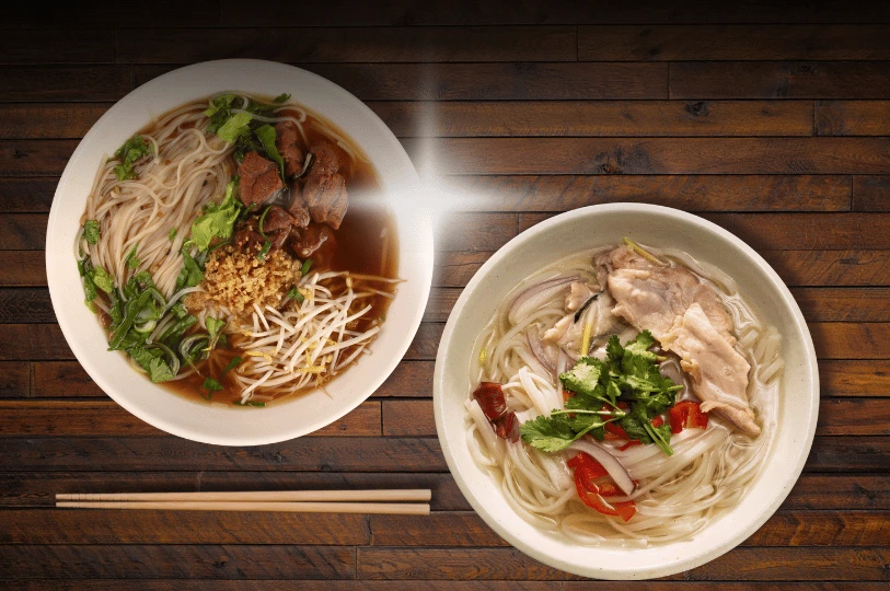 An overhead view of two different bowls of pho on a wooden table, contrasting a dark, rich beef pho with a lighter, clear chicken pho.