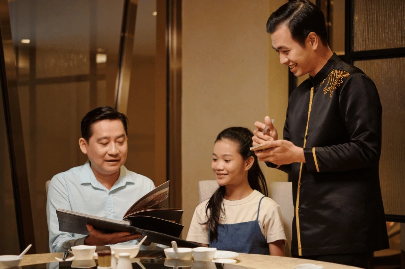 A father and daughter looking at a menu and ordering their meal from a smiling waiter at a restaurant.