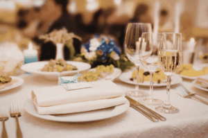 A close-up of an elegant fine dining table setting featuring crisp white linens, crystal wine glasses, a personalized place card, and warm candlelight.