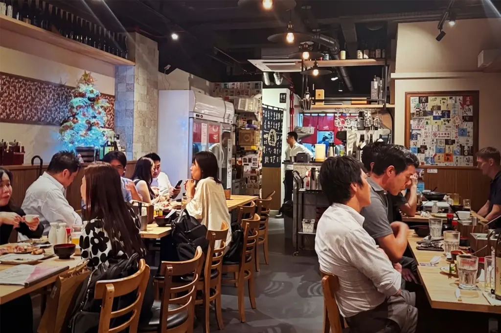 Bustling indoor dining room at Niningashi Japanese Izakaya restaurant on East Coast, showing patrons sitting at wooden tables enjoying food and drinks in a cozy, authentic atmosphere.
