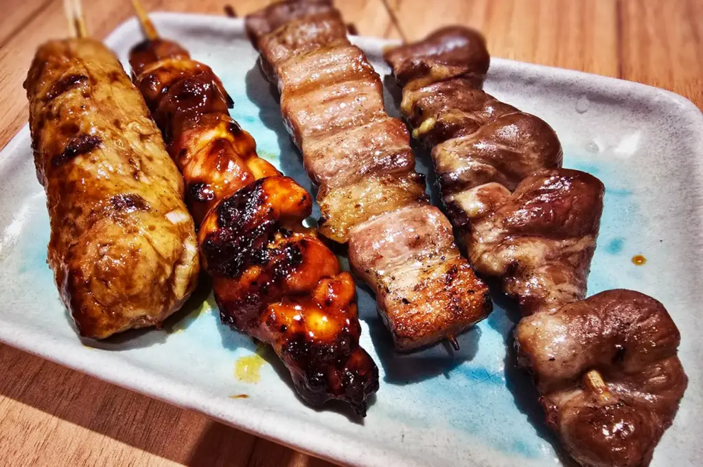 Assorted Japanese yakitori skewers at Niningashi Izakaya, featuring grilled pork belly, glazed chicken Tsukune, and chicken thigh on a ceramic plate.
