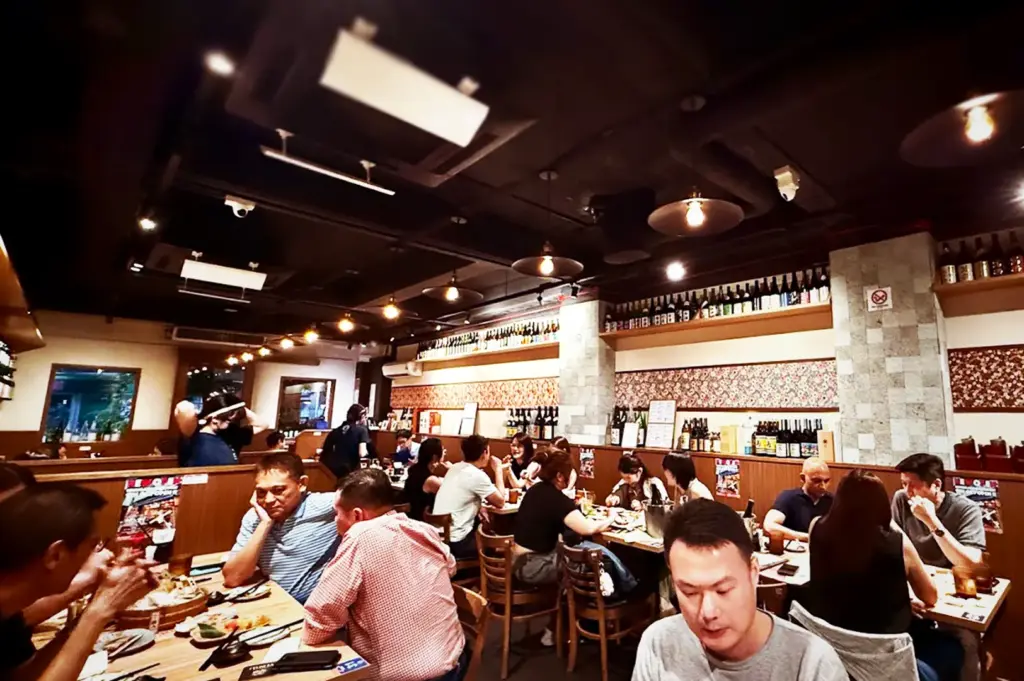 Wide-angle view of the crowded indoor dining area at Niningashi Japanese Izakaya restaurant on East Coast, showing patrons at wooden tables under warm pendant lighting surrounded by sake bottle displays.