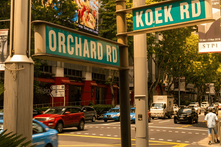 Green street signs for Orchard Road and Koek Road at a sunny intersection, marking the street-level location near Cuppage Plaza.