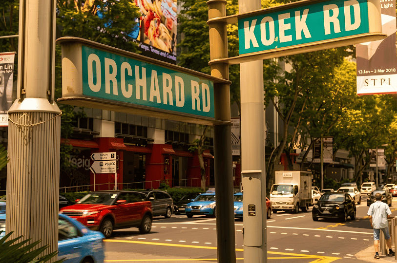 Green street signs for Orchard Road and Koek Road at a sunny intersection, marking the street-level location near Cuppage Plaza.