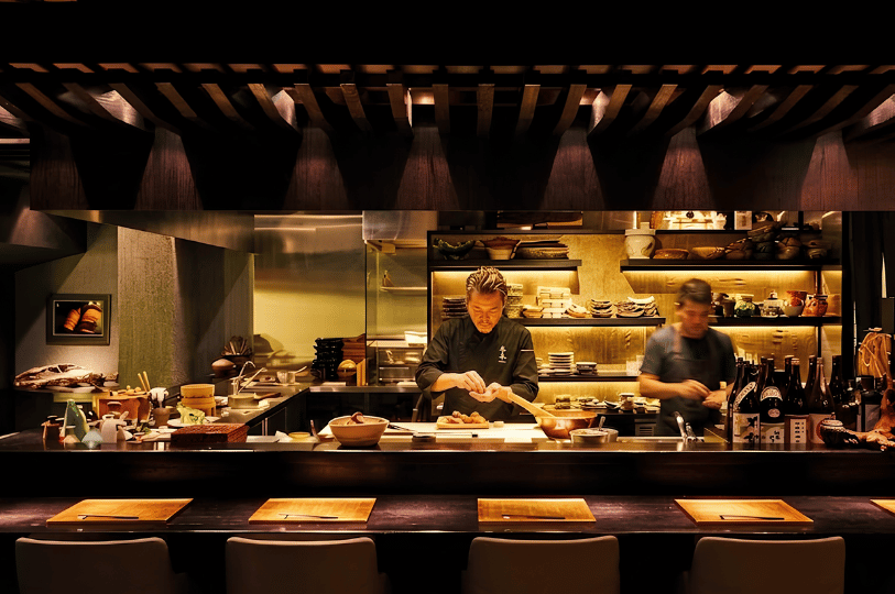 Two chefs carefully preparing food behind a wooden counter in a warm, dimly lit Japanese restaurant, with shelves displaying traditional ceramics and bottles in the background.