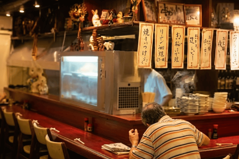 A cozy, authentic Japanese izakaya interior featuring an older patron in a striped shirt dining quietly at a red counter, with handwritten Japanese paper menus hanging above.