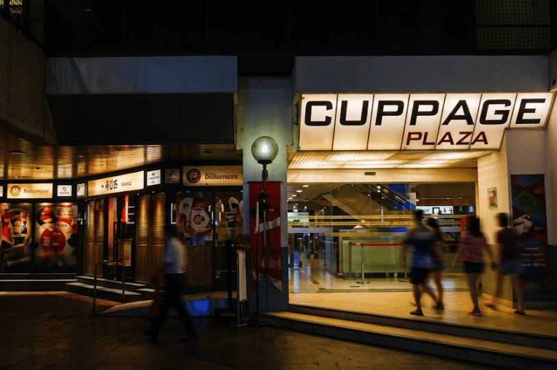 The illuminated exterior entrance of Cuppage Plaza at night, showing the building's signage above glass doors as pedestrians walk past.