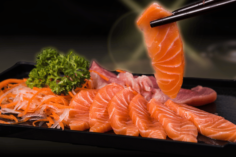 A close-up shot of a freshly prepared sashimi platter featuring neatly arranged slices of raw salmon, shredded carrots, and parsley. A pair of black chopsticks holds up a single, glistening slice of salmon sashimi, emphasizing the food's fresh quality.