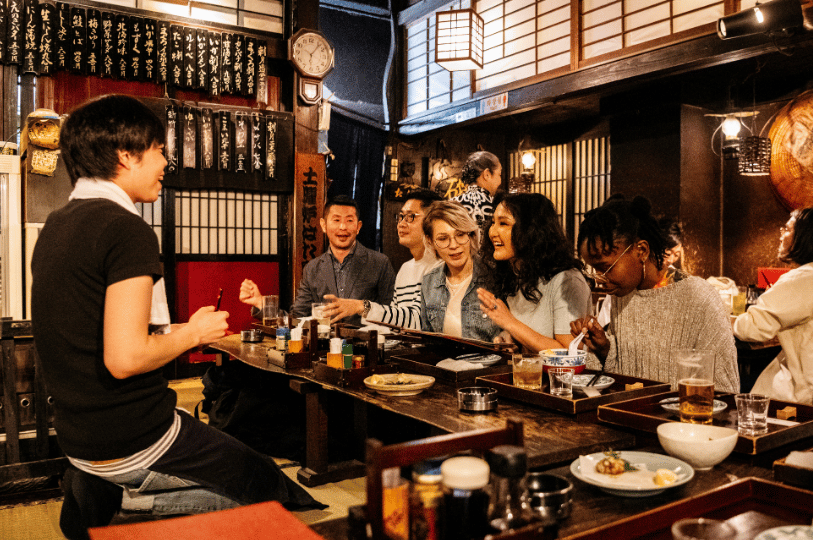 A cheerful, diverse group of friends gathers around a rustic wooden table in a warmly lit, bustling Japanese izakaya. A smiling server kneels to take their order, with traditional Japanese wooden menu boards decorating the background walls.