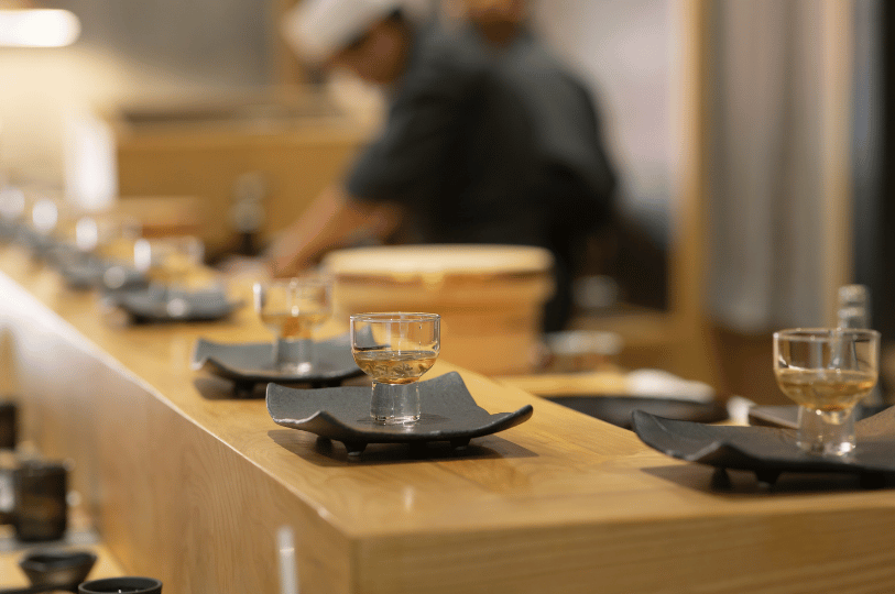Small, clear glass sake cups rest elegantly on dark square coasters along a smooth, light-wood dining counter. In the softly blurred background, a chef is quietly at work, capturing the serene craft of traditional Japanese dining.