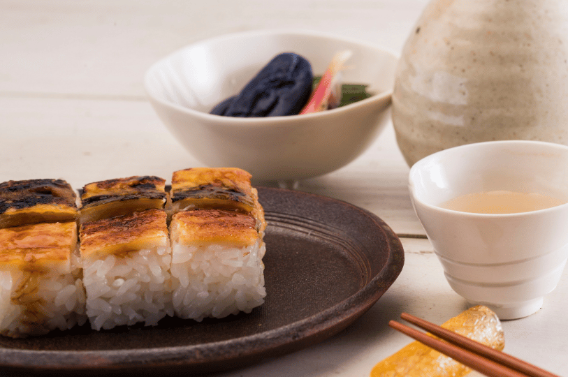 Six pieces of pressed oshizushi, topped with beautifully glazed and seared fish, are served on a rustic dark brown plate. The meal is accompanied by a small white sake cup, a ceramic carafe, and chopsticks resting gently on a traditional stand.