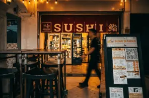 The warmly lit exterior of Sushi Morinaga at night, featuring a red noren curtain with "SUSHI" written across it, glass display fridges, and an outdoor menu board.