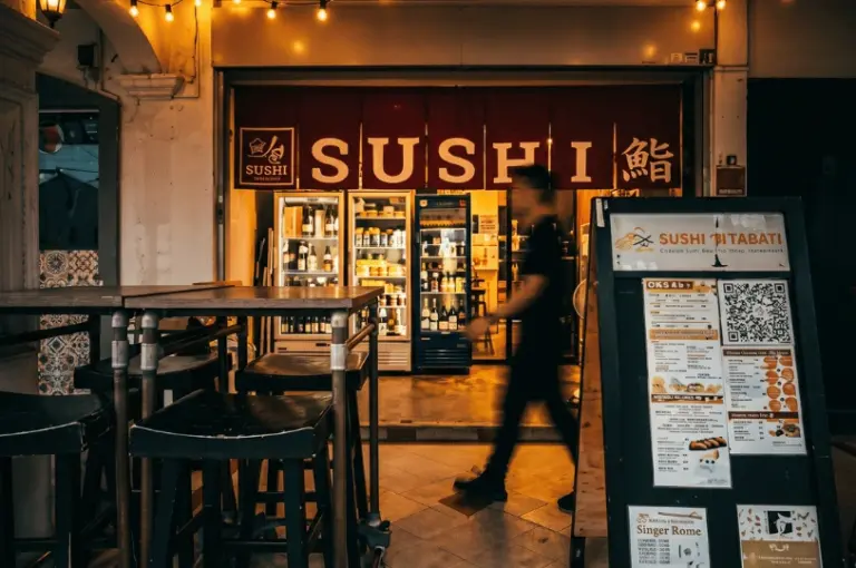 The warmly lit exterior of Sushi Morinaga at night, featuring a red noren curtain with "SUSHI" written across it, glass display fridges, and an outdoor menu board.