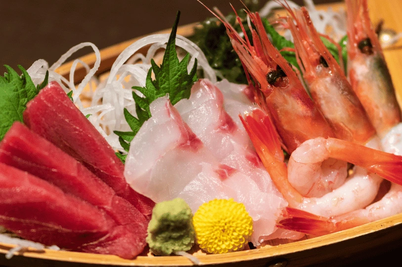 A close-up of a fresh sashimi platter served in a bamboo basket, featuring thick slices of raw tuna, white fish, and whole sweet shrimp.