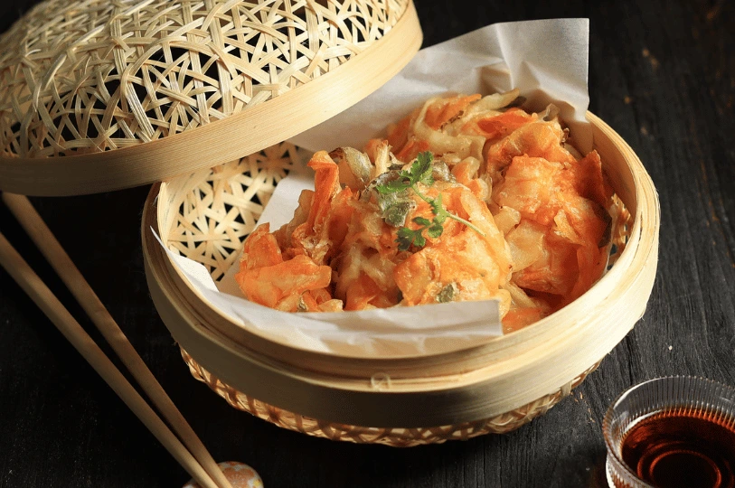 Golden, crispy mixed vegetable tempura served on white paper inside a woven bamboo steamer basket on a dark wooden table.