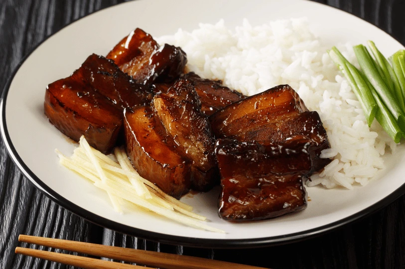 A plate of glistening, dark-glazed braised pork belly served alongside a mound of white rice, shredded ginger, and sliced green onions.