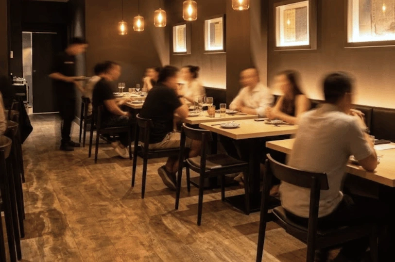 An atmospheric, slightly blurred view of the busy main dining area at Sushi Morinaga, with patrons enjoying their meals at wooden tables under warm pendant lights.