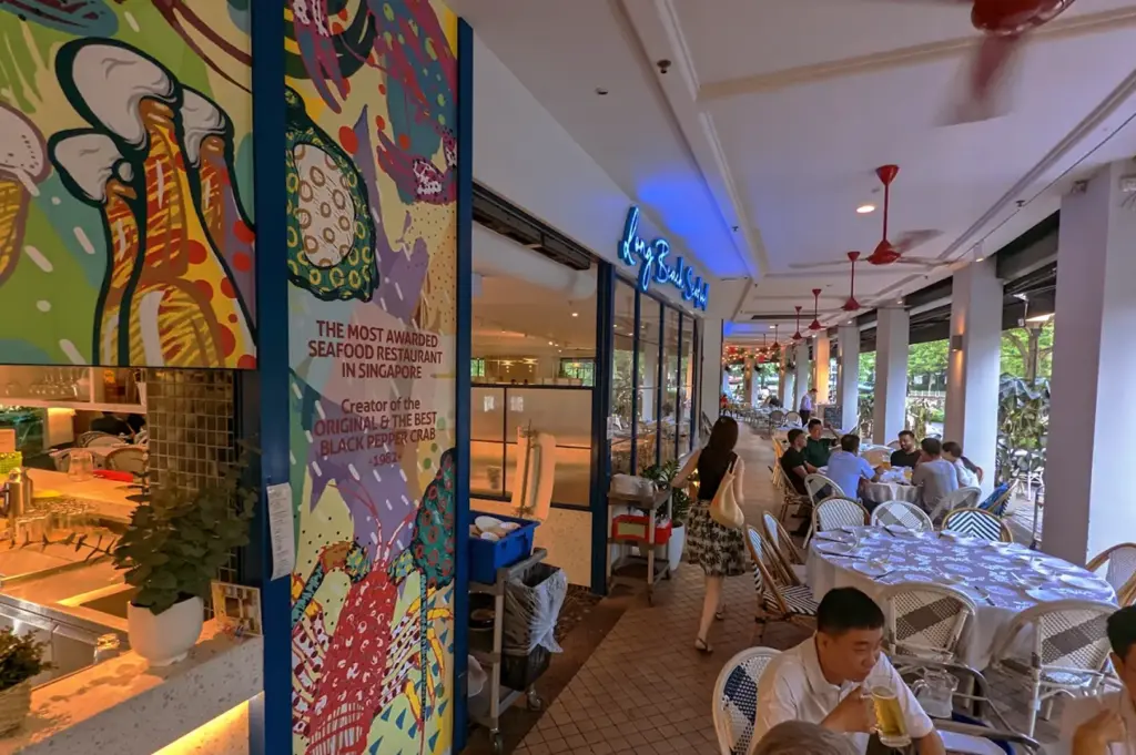 The entrance to Long Beach Seafood restaurant in Singapore, featuring vibrant wall art celebrating their award-winning original black pepper crab of the year, with outdoor dining tables visible.