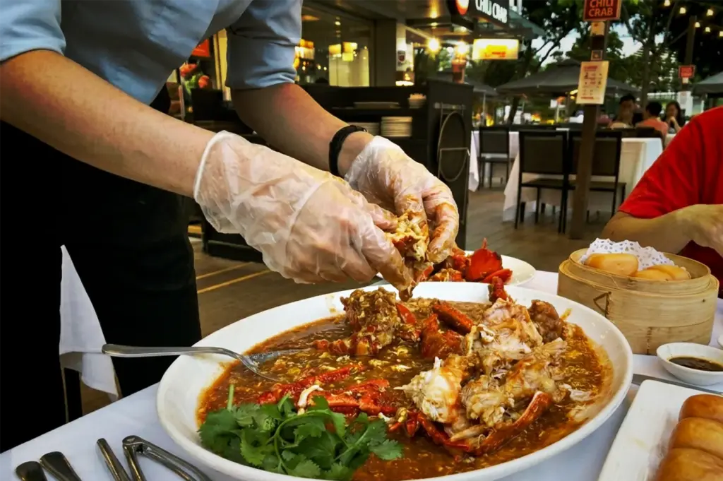 A server wearing plastic gloves deshelling a large plate of Singapore chili crab served with fried mantou buns at an outdoor table.