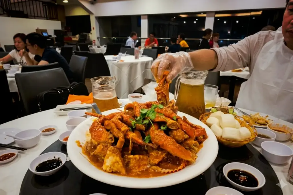 A diner wearing a plastic glove reaches for a platter of Singapore chilli crab at a restaurant table, served with fried mantou buns, dipping sauces, and pitchers of beer in Jumbo Seafood.