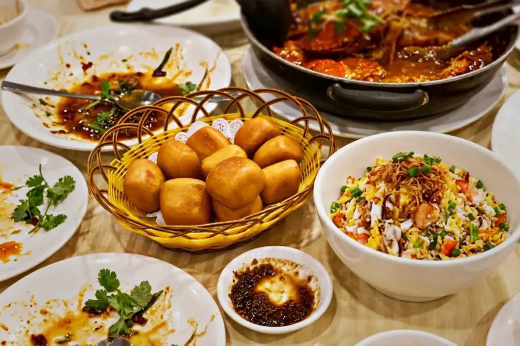 A pan of Singapore chili crab served with fried mantou buns, seafood fried rice, and dipping sauces on a restaurant table in Roland Restaurant.