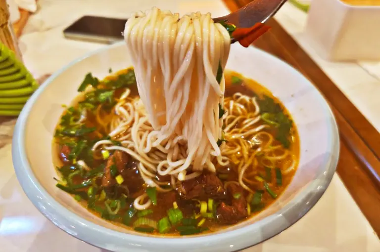 Close-up of thin, springy Chinese noodles being lifted from a savory, dark broth with tender meat and fresh green onions.