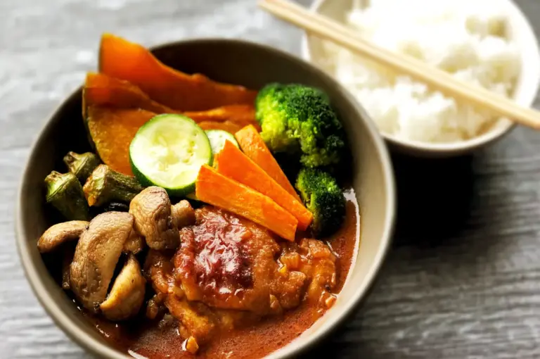 A hearty bowl of Japanese soup curry featuring tender chicken, roasted vegetables like broccoli and pumpkin, and a side of steamed white rice, capturing the light yet flavorful Hokkaido-born specialty.
