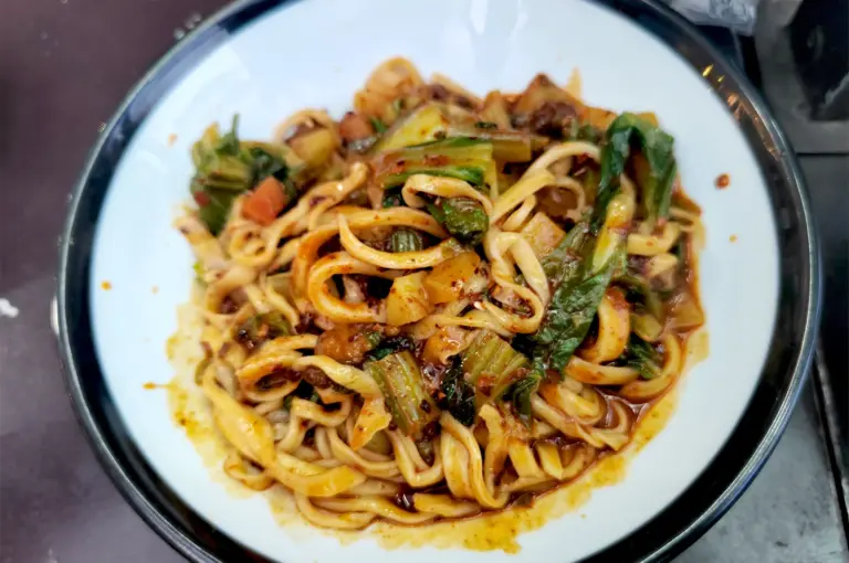 A close-up of a spicy bowl of Chinese wheat-based noodles tossed with fresh greens and savory sauce, showcasing the texture that defines a perfect noodle dish.