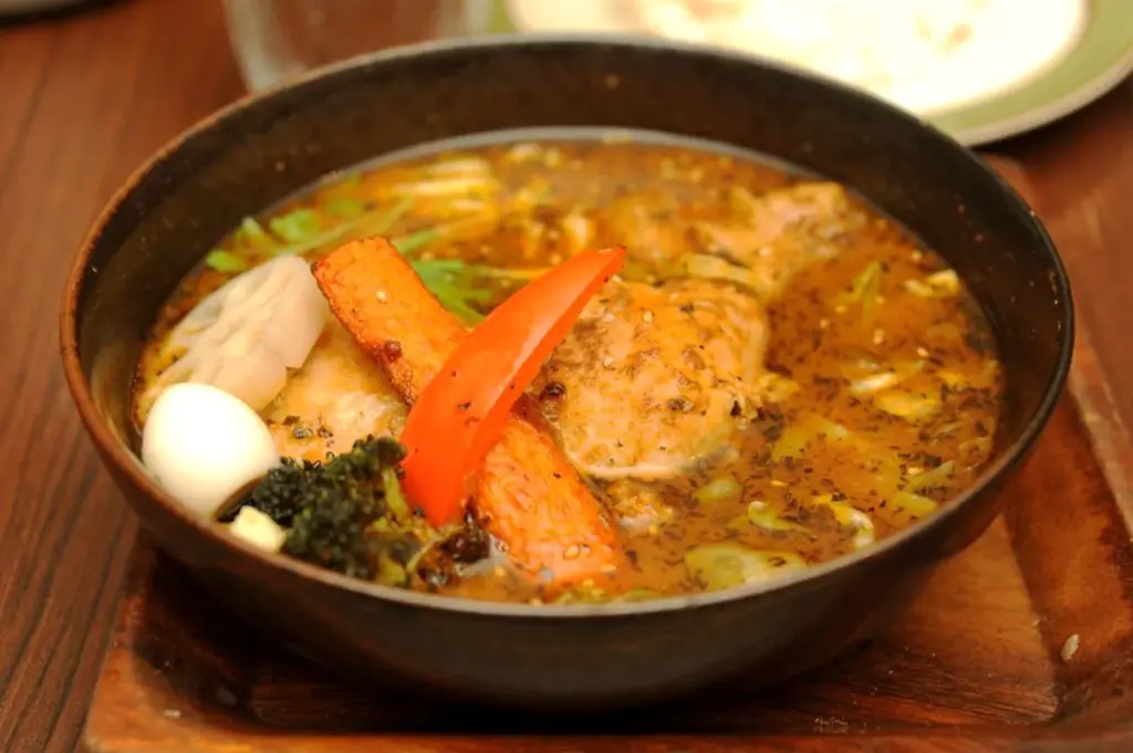 A vibrant close-up of a Japanese soup curry bowl, showcasing a rich, aromatic broth filled with a whole piece of chicken leg, colorful eggplant, bell peppers, and lotus root.