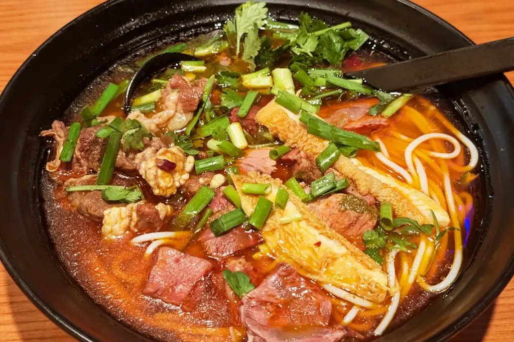 Spicy Sichuan-style Chinese beef noodle soup in a black bowl, featuring thin noodles, tender beef, tripe, and chopped scallions in a vibrant, chili-oil broth.