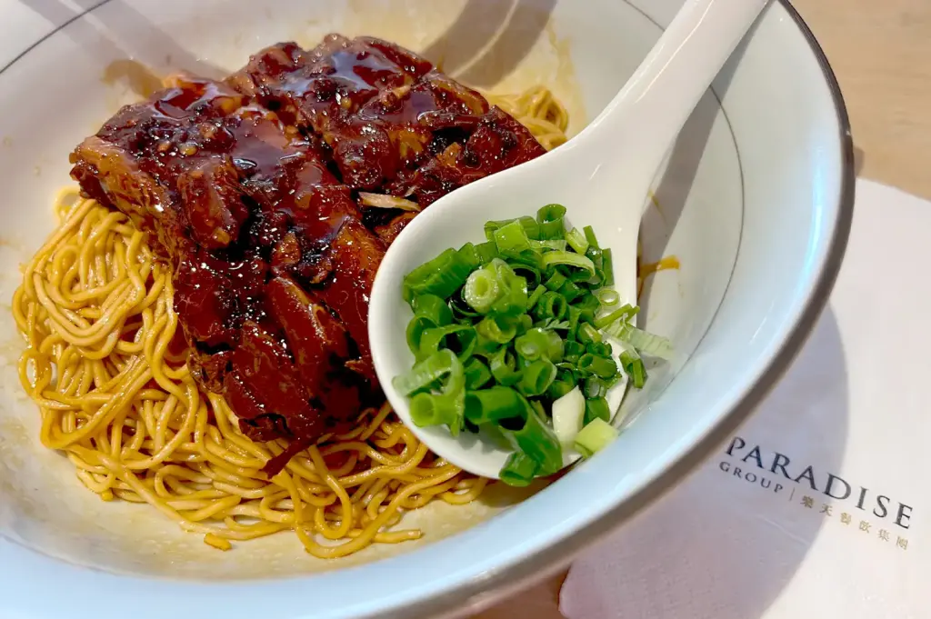 A close-up of a signature dry noodle dish at LeNu Chef Wai's Noodle Bar, topped with tender braised pork belly and served with a side of fresh, vibrant scallions.