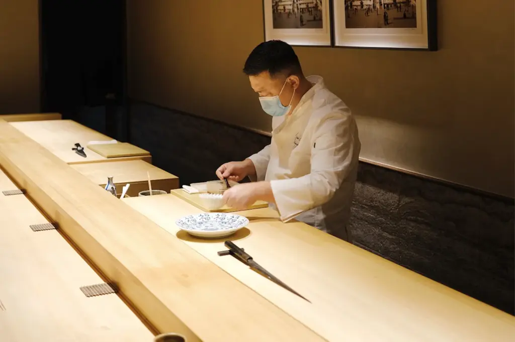 Chef E Ran at Sushi Zen Singapore expertly preparing a meal behind a traditional wooden counter, highlighting the artisanal precision and focus of the restaurant's Omakase service.