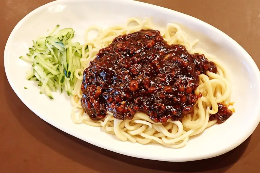 A serving of Zha Jiang Mian, featuring thick Chinese fresh noodles with a chewy texture topped with a rich, savory soybean meat sauce and served with fresh cucumber strips.