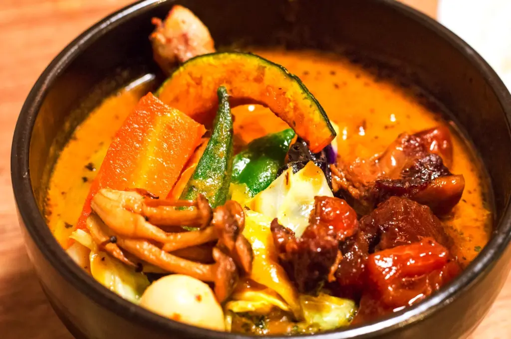 A deep bowl of spicy Japanese soup curry served in a restaurant, packed with braised meat, okra, pumpkin, and mushrooms, illustrating the warm and comforting essence of this Hokkaido dish.