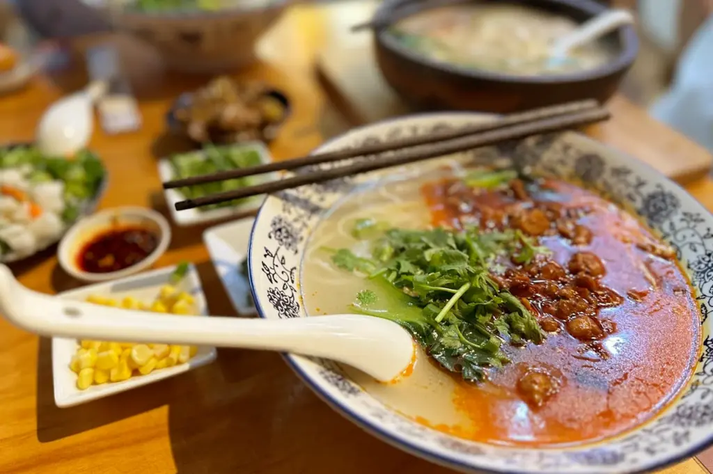 A delicious steaming bowl of Chinese noodle soup served with chili-infused broth, greens, and cilantro, accompanied by other seasonings to bring comforting flavors to the dish.