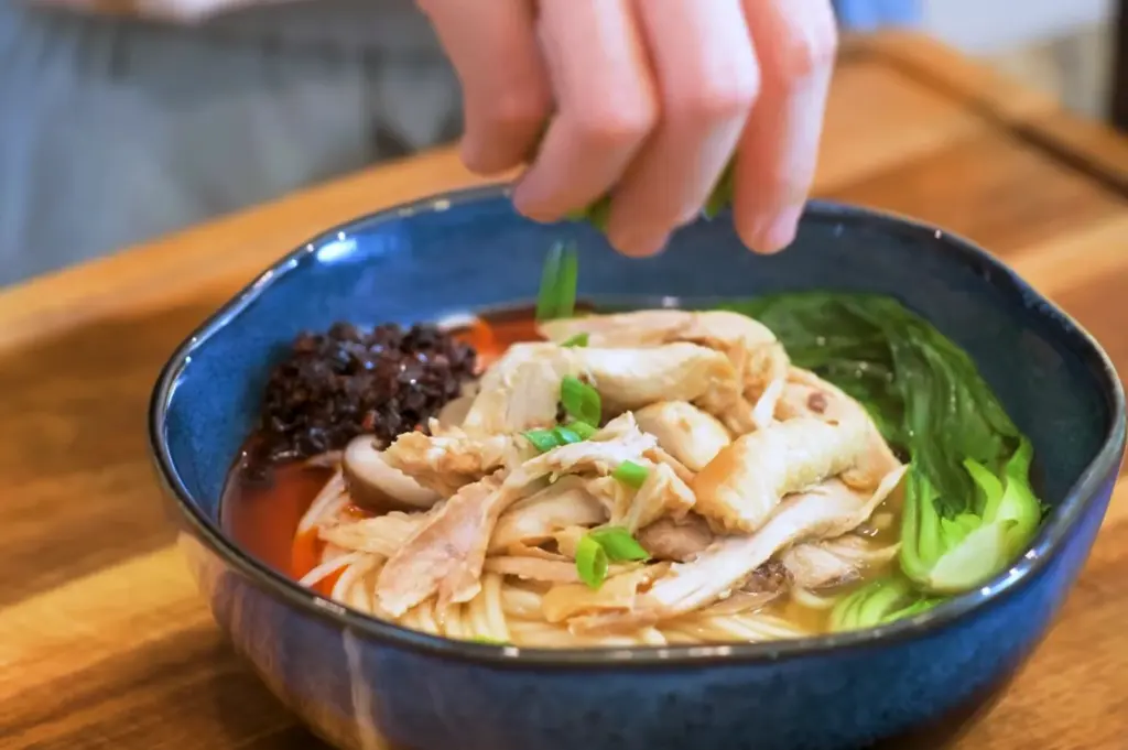 A bowl of homemade chicken noodle soup, a staple food for many, with fresh greens and a dollop of savory black bean sauce being topped with fresh green onions.