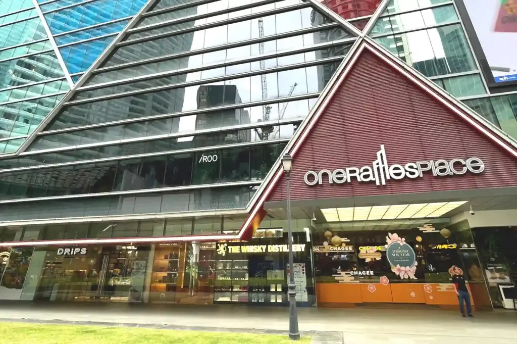 Exterior facade of One Raffles Place in Singapore, featuring modern glass architecture, street-level shops, and the building's signature red triangular entrance sign.