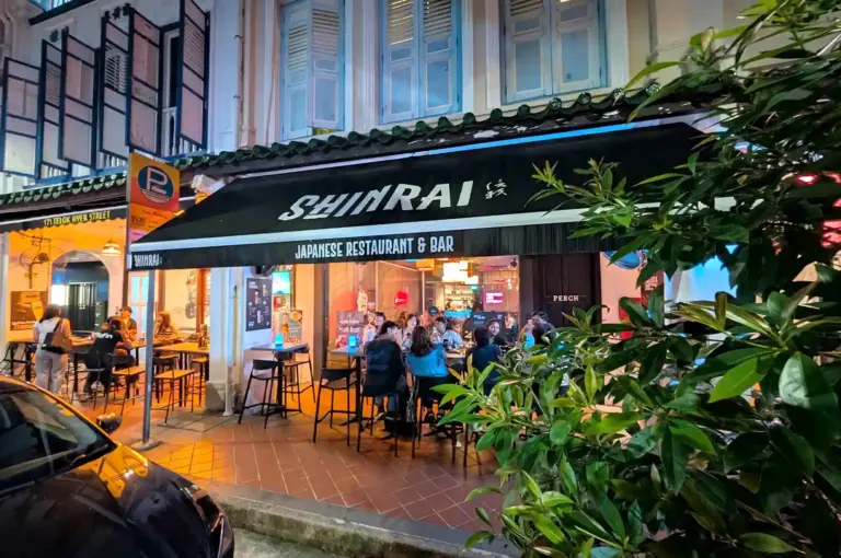 The street-facing storefront of Shinrai Japanese Restaurant & Bar in Singapore, featuring a black awning, outdoor dining tables, and lively evening ambiance.