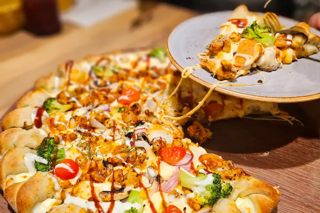 A close-up view of a Korean-style pizza with a stuffed crust, topped with chicken, vegetables, and melted cheese, as a slice is being lifted onto a plate.