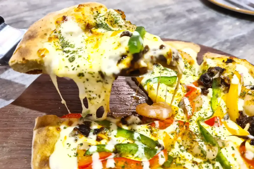 A freshly sliced Korean pizza with green tea signature dough loaded with melted cheese, prawns, and bell peppers, with a piece being pulled away to show long cheese pulls.