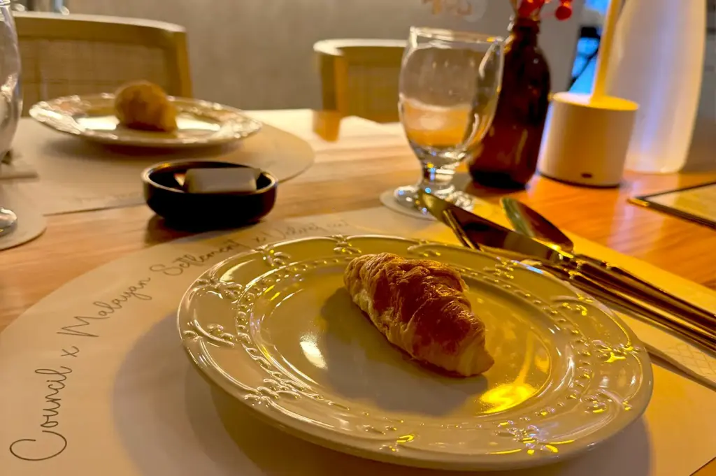 A close-up of a freshly baked croissant on an elegant white plate set on a cozy restaurant table with warm ambient lighting, perfect for slow mornings and quiet reflection.