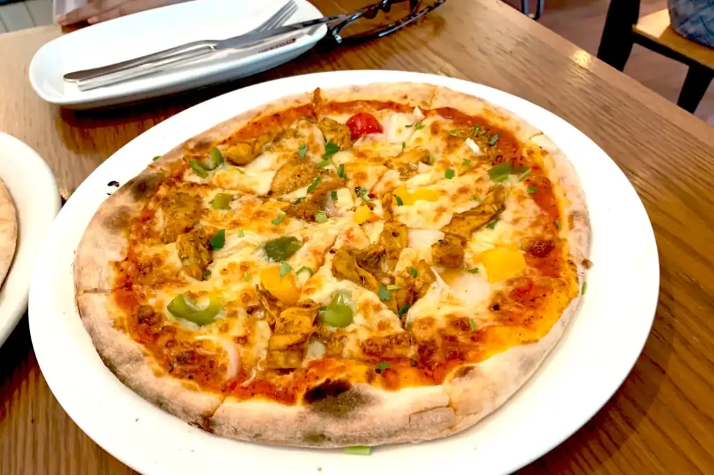 Spizza artisanal Kiara pizza served on a white plate in Singapore, topped with tandoori chicken, bell peppers, melted mozzarella, and fresh herbs.