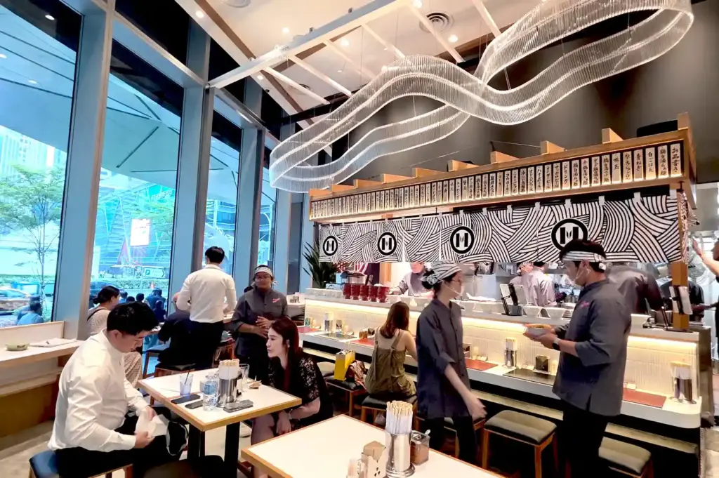 The lively interior of Mensho X ramen restaurant, featuring sleek counter seating, traditional noren curtains, and a striking wavy art installation overhead.