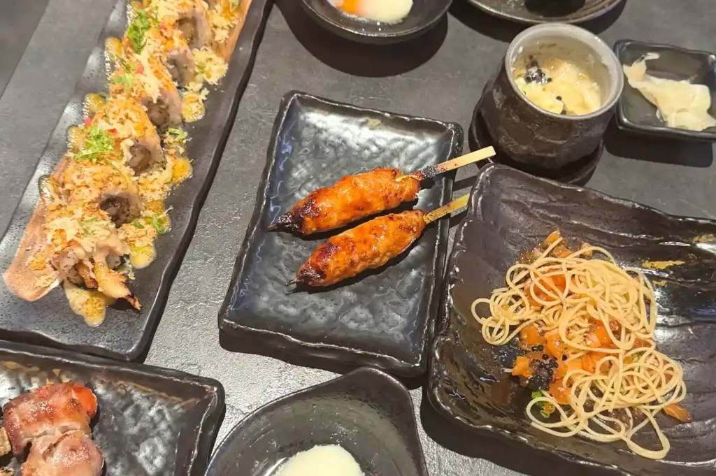 A spread of dishes at Shinrai including sushi rolls, skewered chicken tsukune yakitori, and a plate of cold pasta with roe.