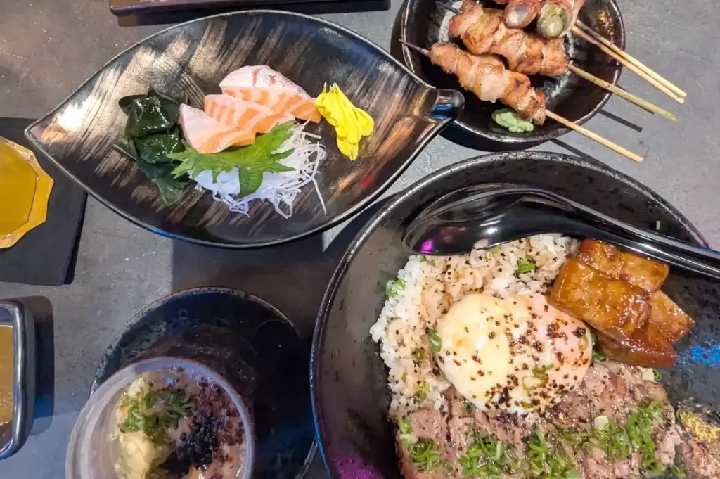 A top-down view of a Japanese spread featuring fresh salmon sashimi slices, skewered meat, and a bowl of rice topped with a soft-boiled egg and glazed meat.