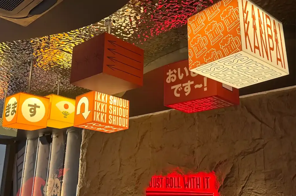 A close-up of colorful illuminated cube lanterns hanging from the ceiling, featuring Japanese typography and playful beer graphics inside Shinrai.