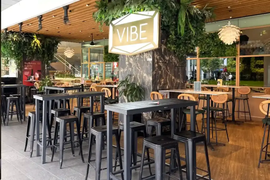 Modern outdoor and indoor high-top seating area featuring lush greenery, wooden ceilings, and an illuminated VIBE sign at VIBE Bistro in Raffles Place.